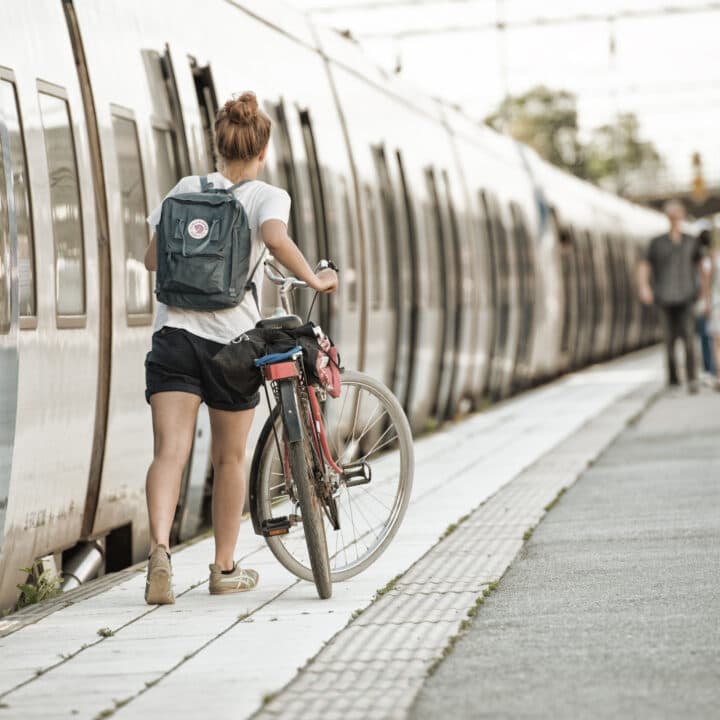 Bilden visar en ung kvinna som står vid en tågplattform med en cykel. Kvinnan är klädd i shorts och en vit t-shirt, och hon har en ryggsäck på ryggen. Hon verkar vara i färd med att parkera eller hämta sin cykel vid sidan av ett tåg. Tåget har en silverfärgad exteriör och plattformen ser ut att vara utomhus. I bakgrunden kan man se andra passagerare som rör sig längs plattformen. Det verkar vara en solig dag och miljön tyder på en urban pendlingssituation.